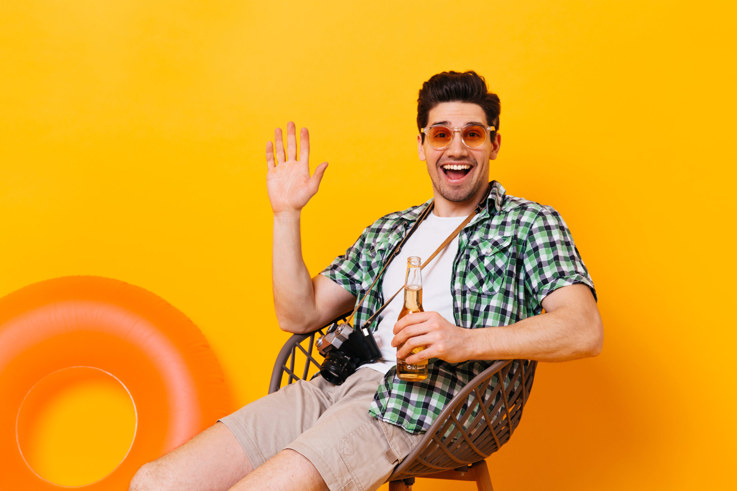 Charming man in green plaid shirt waving his hand, laughing, holding bottle of beer and retro camera on orange background